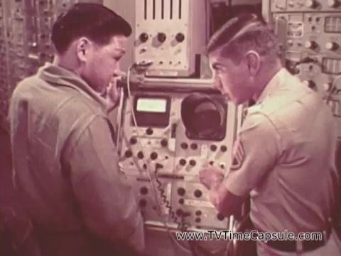 The still from 'Army Recruiting Commercial - Your Man in Waikiki' shows two soldiers interacting in a military setting. One soldier, dressed in a casual uniform, appears to be listening intently while the other, in a more formal uniform, is pointing to equipment or controls. The background features various devices and machinery, suggesting a technical or communication setting. The overall atmosphere reflects a sense of camaraderie and professional engagement within the army.