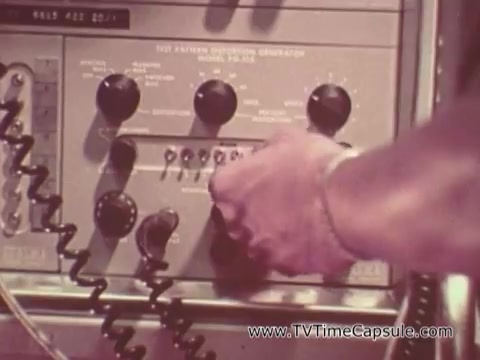 The still from the 'Army Recruiting Commercial - Your Man in Waikiki' depicts a close-up of a person's hand adjusting knobs on a communication device, likely a radio or transmitter. The equipment is vintage, featuring various dials and wires, emphasizing a military or operational context. This scene conveys a sense of action and technology related to Army operations.