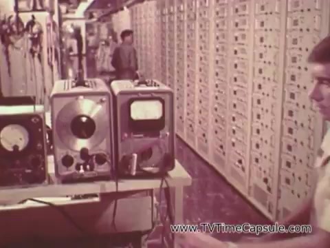 The 16mm still from 'Army Recruiting Commercial - Your Man in Waikiki' features a scene inside a military facility. In the foreground, a young man operates audio-visual equipment, including a reel-to-reel tape recorder and an old television monitor. Behind him, there is a long row of cabinets, suggesting a setting filled with technology and resources likely related to communications or training. The overall atmosphere reflects a structured and organized military environment, emphasizing the theme of technology within the armed forces.
