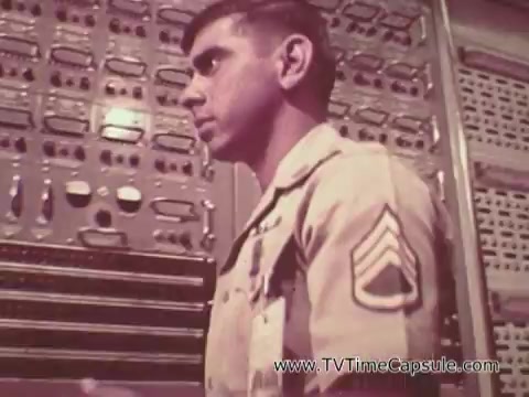 The still from 'Army Recruiting Commercial - Your Man in Waikiki' features a soldier in a military uniform with a rank insignia displayed on his sleeve. He appears engaged in a task or conversation, with various military equipment or control panels visible in the background, suggesting a technical or operational environment. The overall tone conveys a sense of duty and professionalism associated with military service.