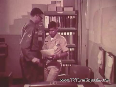 The still from the 'Army Recruiting Commercial - Your Man in Waikiki' shows two soldiers in an office setting. One soldier, wearing a military uniform, is handing a document to the other, who is dressed in a lighter, possibly tropical uniform. The background features filing cabinets and office papers, suggesting a bureaucratic environment. The interaction appears professional, emphasizing a sense of camaraderie and teamwork within the military context.
