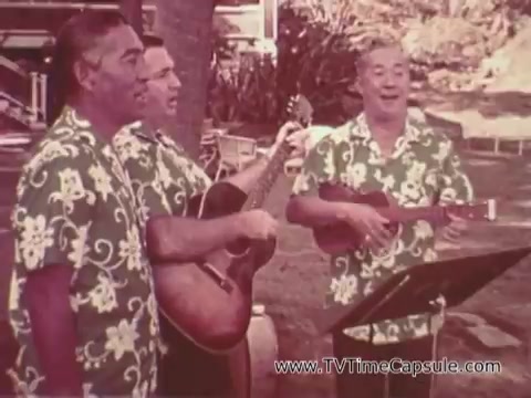 The still from the 'Army Recruiting Commercial - Your Man in Waikiki' features three men dressed in matching floral-patterned Hawaiian shirts. They are performing music, with one playing a guitar and another a ukulele. The setting appears tropical, likely in Waikiki, enhancing the commercial's theme of military service in a vibrant location. The scene conveys a sense of camaraderie and enjoyment, likely aimed at appealing to viewers about joining the Army.