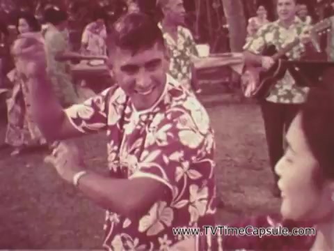 The still from the Army recruiting commercial 'Your Man in Waikiki' features a man in a colorful floral shirt joyfully dancing with a woman, both participating in a lively outdoor Hawaiian event. The background includes other individuals in similar attire, adding to the festive atmosphere. The scene conveys a sense of camaraderie and celebration, promoting a positive image of army life in a tropical setting.