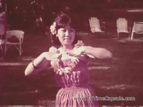 The still from the 'Army Recruiting Commercial - Your Man in Waikiki' features a young woman wearing a purple dress and adorned with leis, performing a dance or gesture with a cheerful expression. The scene is set outdoors, with lawn chairs visible in the background, suggesting a relaxed and festive atmosphere typical of a Hawaiian environment. The overall tone captures a vibrant, inviting vibe associated with the location and the theme of the commercial.