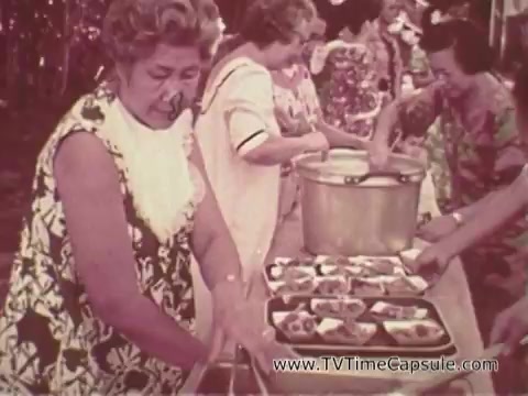 The still features women at a long table serving food, likely in a communal or festive setting typical of a gathering. They wear colorful, floral-patterned dresses, indicative of a tropical or island atmosphere. The scene appears to emphasize community and hospitality, possibly reflecting the cultural backdrop of Waikiki in the context of the Army recruiting commercial. The warm tones and casual ambiance suggest a friendly, inviting environment.