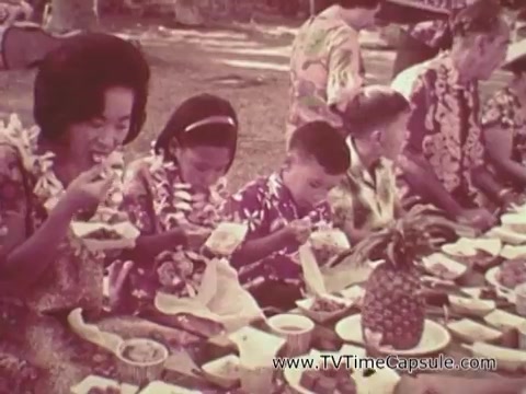 The still from 'Army Recruiting Commercial - Your Man in Waikiki' depicts a family gathering, with people seated around a table filled with various foods. The scene showcases a festive atmosphere, highlighted by participants wearing colorful Hawaiian shirts and leis. A woman and several children are seen enjoying their meal, creating a sense of community and celebration associated with Hawaiian culture. The backdrop suggests a casual outdoor setting, enhancing the spirit of leisure and togetherness.