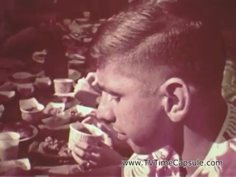 The still shows a young man seated at a table, seemingly in a casual dining setting with various food items scattered around. He is focused on a cup in his hand. The atmosphere appears relaxed and communal, reflecting a moment of camaraderie, likely part of an Army recruiting initiative highlighting life in Waikiki. The color tones suggest a vintage feel typical of 16mm film from that era.