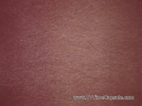 The still from 'Army Recruiting Commercial - Your Man in Waikiki' displays a textured surface with a reddish hue. The image lacks any clear subjects, focusing instead on the background that appears to be fabric or a similar material. The overall tone conveys a vintage aesthetic, characteristic of mid-20th century commercials, aligning with the playful and promotional context of army recruitment in a tropical setting.