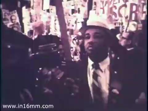 The still features a crowd during a political event, likely a rally or campaign. A man dressed in formal attire, including a suit and hat, is in the foreground, appearing engaged or focused. In the background, numerous placards and signs are visible, emphasizing the political atmosphere of 1968. The overall mood conveys the energy and urgency of an election year, reflecting the social climate of that time.