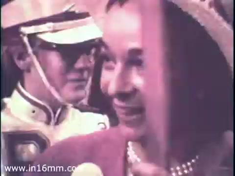 The still from the 1968 PSA for the Chamber of Commerce features a smiling woman holding a flag, likely at a political event or parade. She is dressed elegantly, possibly reflecting the fashion of the era, while a man in a military or ceremonial uniform stands slightly behind her. The image captures the spirit of community engagement and patriotism during the election year. Overall, it conveys a sense of optimism and involvement in the political process.
