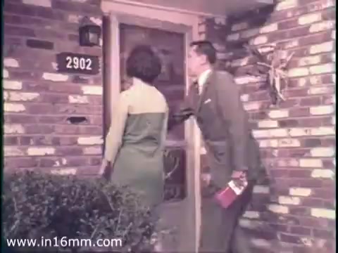 The still depicts a scene from a 1968 PSA featuring a woman and a man approaching a door, likely engaged in political campaigning. The man, dressed in a suit, is knocking on the door while the woman stands beside him, looking toward the entrance. The setting is a suburban neighborhood, characterized by a brick facade and a modest front yard. The overall tone suggests a friendly and approachable atmosphere intended to encourage civic engagement during the election year.