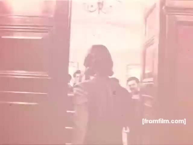 The still from the Souza Cruz Brazilian cigarette commercials of the 1970s shows a character entering through large wooden doors into a well-lit room where several people are gathered. The atmosphere appears social, possibly hinting at a gathering or celebration. The soft focus and muted color tones typical of the era contribute to a nostalgic vibe. This commercial likely aimed to create an image of camaraderie and leisure associated with smoking.