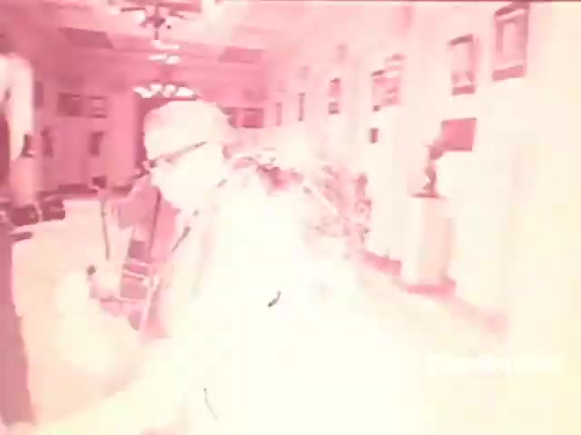 The still depicts a scene from a Souza Cruz cigarette commercial from the 1970s, featuring a bright and slightly washed-out color palette. It shows a gathering, likely in an indoor setting adorned with decor typical of that era. A man, possibly the central figure, is seen wearing glasses and a suit, suggesting a formal context. The lighting and composition convey a nostalgic ambiance, characteristic of vintage advertisements aimed at portraying a lifestyle associated with smoking at that time.