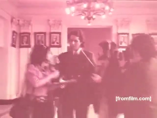 The still depicts a scene from a 1970s Brazilian cigarette commercial for Souza Cruz, featuring two primary figures in a lively, social setting. One individual, dressed in formal attire, appears to engage with a woman in a casual outfit, suggesting a dynamic interaction. The background showcases elegant decor, likely indicating a luxurious venue, with portraits on the walls and a chandelier overhead, reflecting the aesthetic of that era. The overall atmosphere conveys a blend of sophistication and casual conversation typical of advertisement themes from that period.