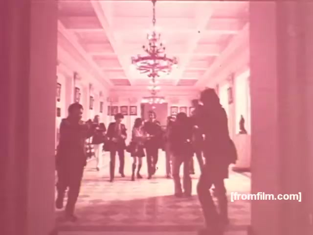 The still depicts a corridor, likely in a formal building, characterized by a chandelier and classical architecture. A group of people is engaged in a lively interaction, suggesting a social or promotional event, typical of the vibrant style of the 1970s Brazilian cigarette commercials. The color palette appears warm, with a slight pink tint, which adds to the nostalgic aesthetic of the era. The scene likely aims to convey a sense of camaraderie and lifestyle associated with the brands Albany and Richmond.