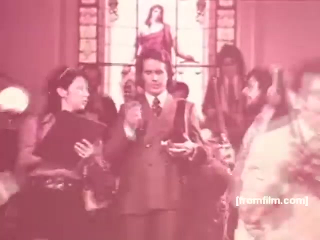 The still depicts a scene from a Souza Cruz cigarette commercial from the 1970s, likely for the Albany or Richmond brand. In the foreground, a well-dressed man is holding a cigarette, surrounded by a lively crowd. To his left, a woman appears engaged, while the background features a stained glass window, possibly indicating a religious or cultural context. The overall aesthetic is vibrant and nostalgic, characteristic of advertising from that era.