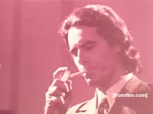 The still features a man in a suit, holding a cigarette close to his lips as he prepares to light it. The scene is characterized by a soft, warm color palette, creating a nostalgic and somewhat glamorous vibe typical of 1970s advertisements. The focus on the act of smoking suggests a sense of sophistication and allure associated with the brand. This visual reflects the marketing strategies of Souza Cruz for brands like Albany and Richmond, aimed at evoking a stylish lifestyle.