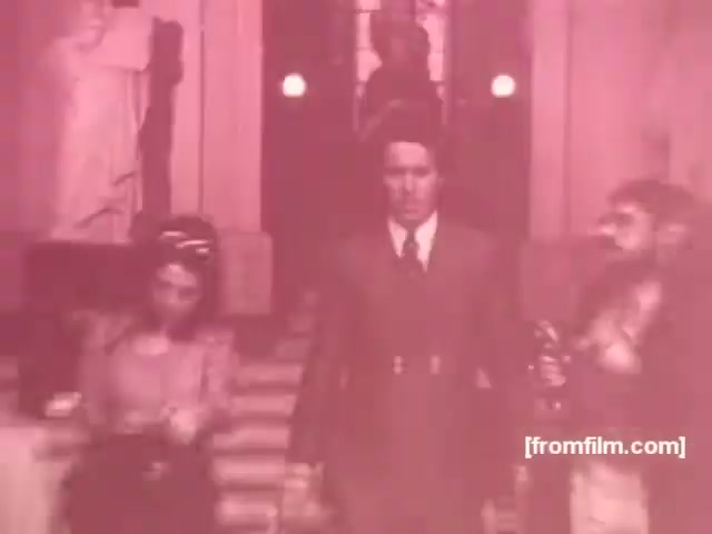 The still features a scene from a 1970s Souza Cruz cigarette commercial. A well-dressed man, likely the main character, walks down a staircase, flanked by two individuals—one woman and one man. The setting appears to be grand, possibly a hall or lobby, with sculptures in the background hinting at an upscale atmosphere. The image has a pinkish tint, typical of the film style from that era, adding a nostalgic quality to the commercial. The overall composition emphasizes sophistication and allure associated with the advertised cigarette brands, Albany and Richmond.
