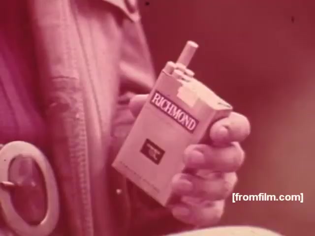 The still shows a close-up of a person's hand holding a pack of Richmond cigarettes, prominently displayed with a simple design. The color palette has a vintage feel, likely due to the film's age, giving it a nostalgic and retro aesthetic. The individual appears to be dressed casually, adding to the relaxed vibe typical of cigarette commercials from the 1970s. Overall, it captures a moment of casual consumption associated with the brand.