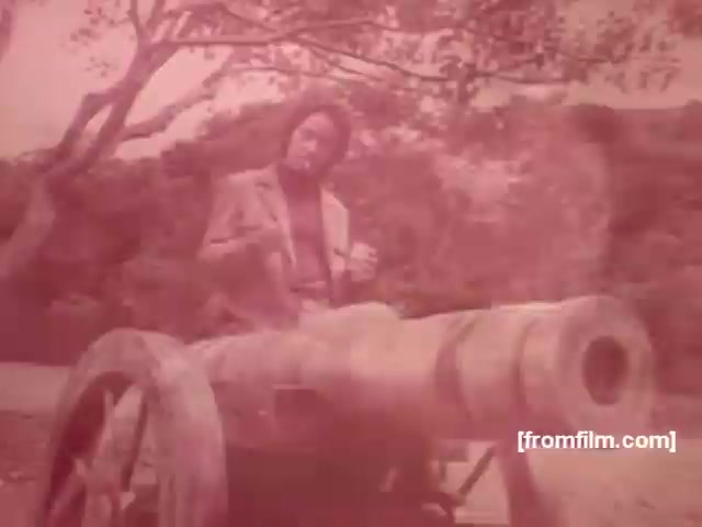 The still captures a scene from a Souza Cruz cigarette commercial, likely from the early 1970s. It features a man dressed in a suit, seated beside a cannon, suggesting a historical or playful context. The sepia tone adds a nostalgic atmosphere, typical of vintage advertisements. This stylistic choice, along with the unusual setting, aims to evoke intrigue and memorable associations with the featured cigarette brands, such as Albany or Richmond.