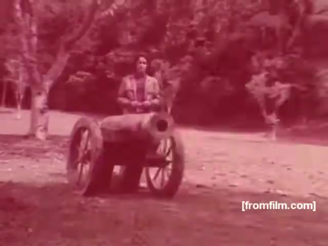 The still from the Souza Cruz Brazilian cigarette commercial features a solitary figure sitting on a historical cannon in a lush, green landscape. The scene has a vintage aesthetic, characterized by a reddish tint that enhances its nostalgic feel. This visual likely aims to connect the brand with themes of strength and historical significance, typical of advertising strategies in the 1970s. The tranquil environment contrasts with the weight of the cannon, suggesting a deeper narrative about tradition and legacy associated with the cigarette brand.