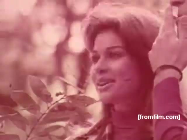 The still features a woman with voluminous hair, smiling while holding a cigarette. She appears in a natural setting, surrounded by foliage, evoking a sense of relaxation and enjoyment. The color tone is pinkish, characteristic of the aesthetic often used in 1970s advertisements. The imagery suggests a blend of lifestyle and product promotion, typical of cigarette commercials from that era, aiming to associate smoking with leisure and sophistication.