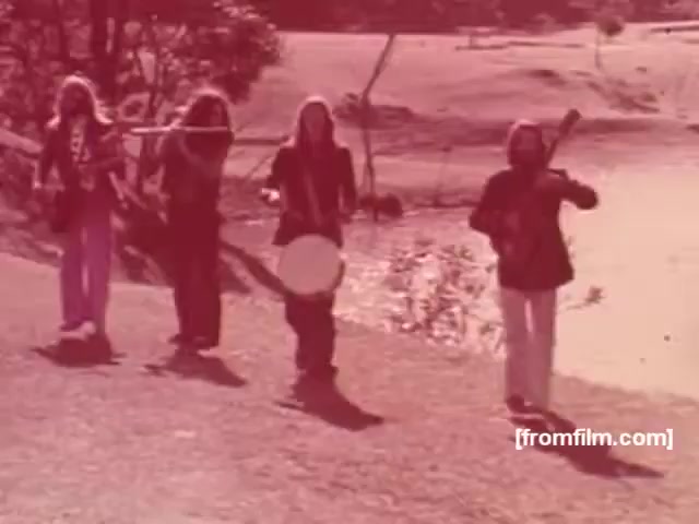 The still from the Souza Cruz Brazilian cigarette commercials features four musicians walking in a natural setting, likely a field or by a body of water. They are dressed in casual, colorful clothing typical of the 1970s, with one playing a flute, another holding a drum, and the others with string instruments. The scene conveys a sense of camaraderie and outdoor enjoyment, aligning with the carefree, vibrant aesthetic of the era. The warm, slightly faded color palette adds a nostalgic feel to the image.