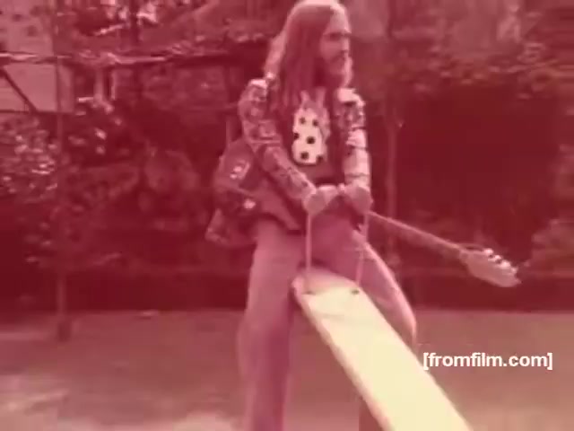 The still features a man with long hair sitting on a seesaw, playing an electric guitar. His attire reflects the 1970s fashion, sporting a patterned shirt with vibrant colors. The overall tone of the image has a warm reddish hue, typical of film from that era. The setting appears to be a backyard or garden, contributing to a laid-back and casual vibe associated with the era's youth culture. This scene likely aims to convey a sense of freedom and enjoyment, fitting the advertising style of the time for Souza Cruz's cigarette brands like Albany and Richmond.