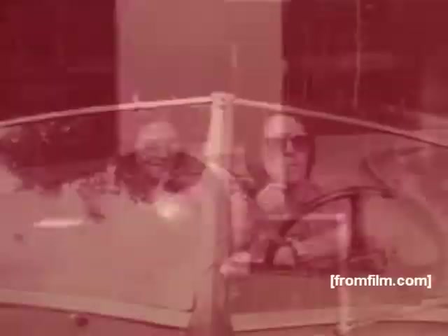 The still features two men in sunglasses driving a convertible, evoking a sense of leisure and sophistication typical of 1970s advertisements. The warm, reddish tint adds a nostalgic atmosphere, which is common in vintage cigarette commercials. The scene suggests a lifestyle of enjoyment and freedom, aligning with the messages of both Albany and Richmond cigarette brands at the time. The overall composition emphasizes a carefree, glamorous lifestyle associated with smoking in that era.