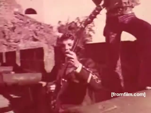 The still from the Souza Cruz cigarette commercials from the 1970s features a dramatic scene. A male figure, presumably an actor, is holding a guitar and is dressed in a military-style outfit, suggesting a sense of action or adventure. The backdrop includes a rugged, urban environment, possibly symbolizing the era's themes of rebellion or youth culture. This visual style reflects the bold marketing strategies typical of cigarette advertisements during that time, aiming to evoke emotions tied to freedom and masculinity, often associated with rock music and counterculture.