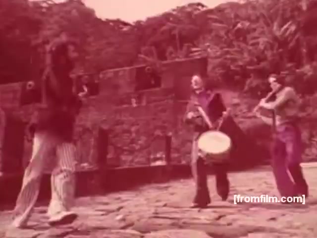 The still features a vibrant outdoor scene from a 1970s Brazilian cigarette commercial. It shows three musicians engaged in a lively performance, with one playing a drum and another a violin. The backdrop includes lush greenery, contributing to a festive atmosphere. The overall aesthetic has a nostalgic feel, typical of that era in advertising, with a focus on cultural expression and community. The warm tones of the image enhance its retro charm.