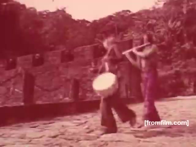 The 16mm still from the Souza Cruz Brazilian cigarette commercials presents a vibrant scene featuring two musicians. One plays a flute while the other beats a drum, set against a backdrop that suggests a natural, possibly rural environment. The color palette leans toward reddish tones, indicative of the era’s aesthetic. This captures a cultural element, likely intended to evoke a sense of tradition and local pride, appealing to viewers during the 1970s in Brazil.