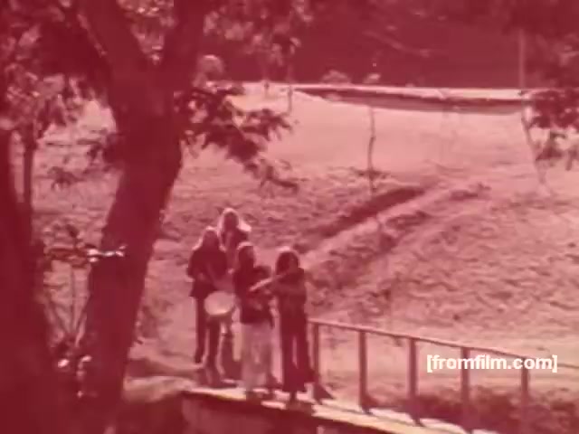 The still from the 1970s Souza Cruz cigarette commercials features a serene outdoor setting, likely in Brazil. In the foreground, several individuals are engaged in a musical activity near a wooden railing, with a clear view of a grassy area behind them. The scene has a warm, nostalgic tone, characteristic of the era's advertising style, emphasizing a laid-back lifestyle associated with smoking. This captures the relaxed and communal spirit promoted by brands like Albany and Richmond during that decade.