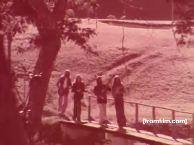 The still depicts a group of four musicians walking along a narrow bridge in a natural setting, likely promoting a sense of freedom and lifestyle associated with smoking. The imagery is bathed in a reddish tint, typical of the era’s aesthetic, evoking a nostalgic and relaxed atmosphere. The surrounding environment appears serene, suggesting an outdoor lifestyle that connects with the brand’s messaging. Overall, the visual captures the cultural vibe of Brazil in the 1970s, aligning with the marketing strategies of Souza Cruz at the time.
