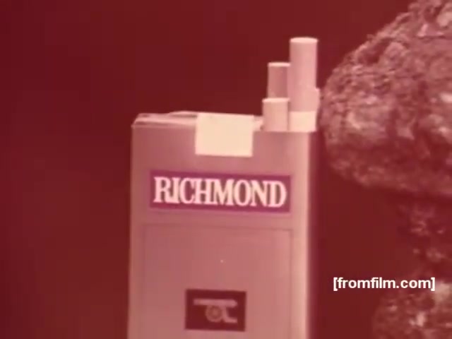 The 16mm still features a pack of Richmond cigarettes prominently displayed against a muted, possibly reddish background. The pack has a sleek, minimalist design, showcasing the brand name 'RICHMOND' in bold, white letters. Above it, a few cigarettes are partially visible, suggesting the product's appeal. The overall aesthetic reflects the vintage style of 1970s advertising, characterized by a focus on simplicity and striking visuals.