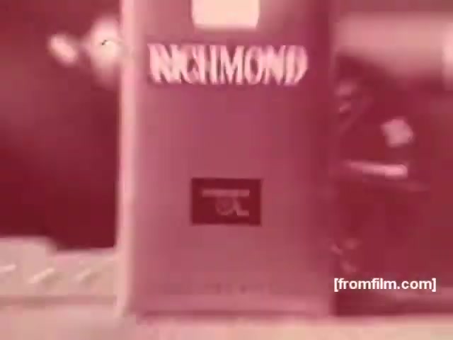 The still features a close-up of a Richmond cigarette package, characterized by its bold graphics and branding typical of 1970s cigarette advertising. The color palette is muted, suggesting an aged or vintage quality. In the background, there appears to be a glimpse of imagery or design, likely related to the lifestyle marketed by the commercial. The focus remains on the product, highlighting its branding and appeal during that era.