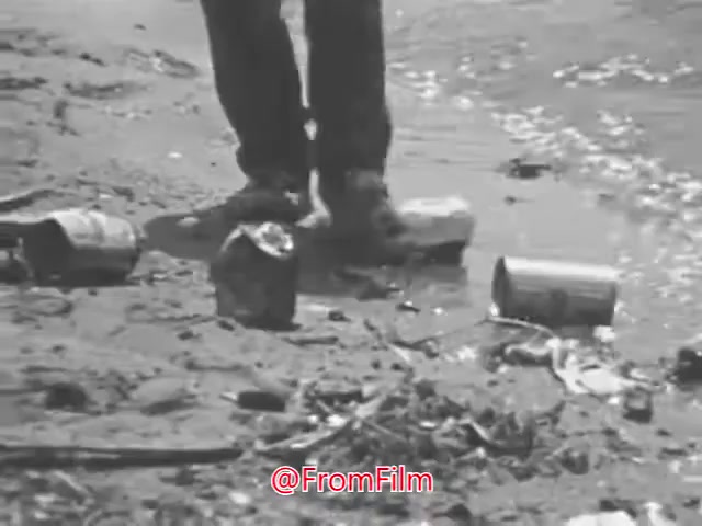 The still from the 'Department of Interior Stop Pollution PSA - 1960's' features a close-up of a person's legs standing in shallow water, surrounded by litter, including cans and debris. The image starkly contrasts with the cheerful, idyllic lyrics about clear days and joyful outdoor activities, highlighting the negative impact of pollution on natural environments. The scene embodies a call to awareness, emphasizing the importance of preserving clean waterways for the enjoyment of future generations.