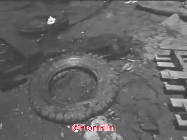 The still from the 1960s PSA highlights environmental pollution, presenting a scene cluttered with debris, including a discarded tire and various scraps. The contrast between the upbeat lyrics about clear days and joyful water activities and the dismal image of pollution underscores a poignant message about the impact of waste on natural spaces. This visual critique serves to raise awareness about the importance of keeping waterways clean.