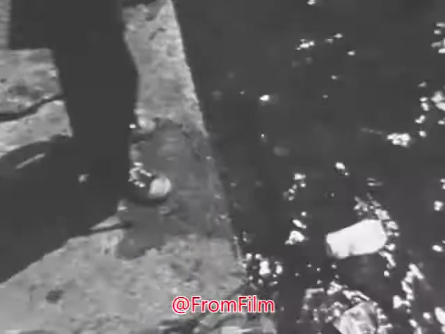 This 16mm still from the 'Department of Interior Stop Pollution PSA - 1960's' depicts a person standing beside a waterway. The focus is on their feet and the edge of a concrete bank, where litter—specifically a plastic bottle—can be seen floating in the water. The image conveys a contrast between the intended message of enjoying clean natural environments and the reality of pollution in waterways, aligning with the PSA's themes of environmental awareness and conservation.