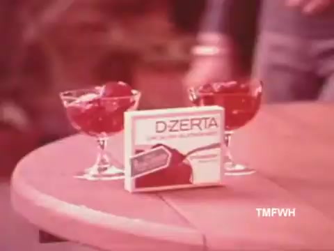 The still from the D-Zerta TV commercial showcases a simple setup featuring two elegant dessert bowls containing jello and a prominently displayed box of D-Zerta on a wooden table. The scene exudes a nostalgic 1970s-1980s aesthetic, with warm colors and a casual, inviting atmosphere typically associated with vintage food advertisements. This product was aimed at consumers seeking a sugar-free gelatin dessert alternative.