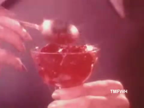The still from the D-Zerta TV commercial features a close-up of a hand gracefully holding a glass dish filled with red gelatin. A spoon is poised above the gelatin, suggesting a moment of indulgence. The lighting has a soft, warm glow, emphasizing the vibrant color of the dessert, typical of the promotional style of the 1970s-1980s. The overall presentation evokes a sense of elegance and appeal, in line with the product's positioning before being replaced by Sugar-Free Jell-O.