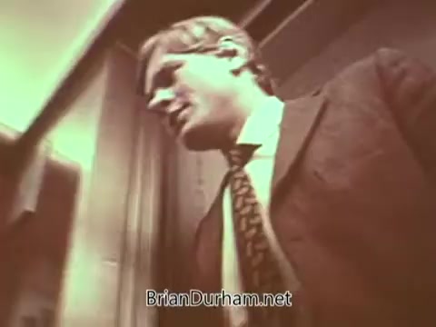 The still from the 'Radio Free Europe PSA' features a man in a suit, appearing to exit an elevator or a confined space. The image has a warm, vintage tint and captures a moment that conveys a sense of urgency or purpose. The man's expression suggests he may be engaged in a serious discussion or activity related to the mission of Radio Free Europe, emphasizing themes of communication and information dissemination. The focus appears to be on the importance of free speech and access to information during a time of political tension.