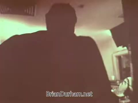 The still from 'Radio Free Europe PSA' features a shadowy figure, likely a person, in a dimly lit interior space. The focus appears to be on the individual’s silhouette, creating a mysterious atmosphere. Surrounding details suggest a setting related to broadcasting or communication, possibly emphasizing the theme of the PSA.