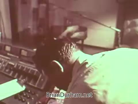 The still shows a person focused on operating a soundboard in a studio setting, presumably for a Public Service Announcement (PSA) related to Radio Free Europe. The atmosphere suggests a vintage broadcast environment, with equipment and microphones visible, emphasizing the importance of media communication during that era. The image captures the dedication of those involved in delivering impactful messages through radio.