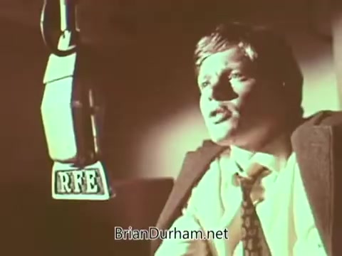 The still depicts a man speaking into a microphone labeled 'RFE,' likely representing Radio Free Europe. He appears earnest and engaged, possibly delivering a message or announcement. The lighting casts a warm tone, contributing to a serious atmosphere typical of public service announcements. The focus on the speaker suggests a direct appeal to the audience regarding important issues.