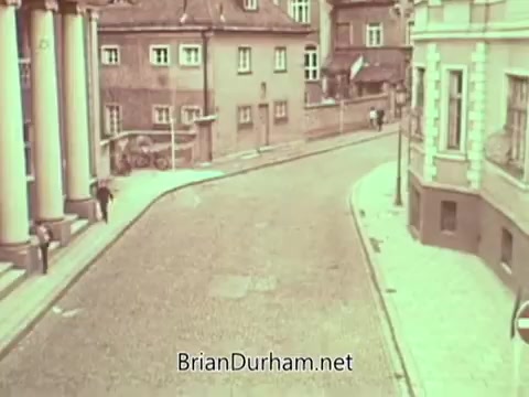 The still features a street scene depicted in a muted color palette, showcasing a cobblestone road winding between buildings with a historical architectural style. One person walks along the sidewalk, contributing to a sense of quiet urban life. The overall atmosphere seems reflective and perhaps nostalgic, typical of public service announcements aimed at conveying important messages.