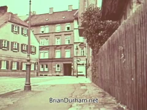 The still from 'Radio Free Europe PSA' depicts a quiet street scene characterized by older, multi-story buildings with a slightly muted color palette. The architecture suggests a European setting, with a lamppost on the left and a wooden fence lining the right. The overall mood appears somber, possibly reflecting the historical context of the PSA's message around freedom and communication during the Cold War era.