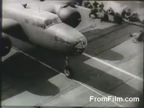 The still depicts a B-25 bomber closely positioned on the deck of the USS Hornet, preparing for takeoff during the Doolittle Raid on April 18, 1942. Crew members are visible in the background, indicating the urgency and teamwork involved in this historic mission. The black-and-white footage captures the intensity and significance of the moment, highlighting a pivotal event in World War II.