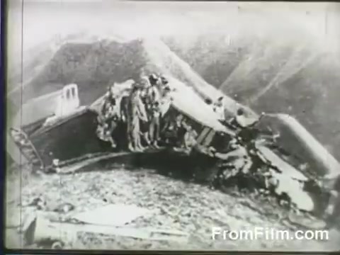 The still depicts a damaged B-25 aircraft, likely from the Doolittle Raid, with a group of individuals inspecting the wreckage. The scene is set against mountainous terrain, indicative of the challenging conditions faced by the raiders following their historic mission launching from the USS Hornet on April 18, 1942. The imagery captures the aftermath of their daring strike, highlighting the perils encountered during the operation.
