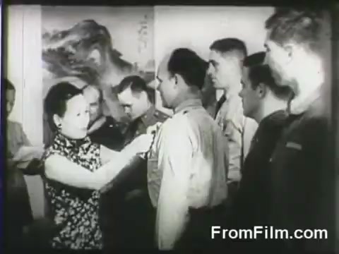 The still depicts a ceremony where a woman, dressed in a traditional Chinese outfit, is pinning a medal onto a soldier, likely a Doolittle Raider. Surrounding them are several military personnel, observing the event with a backdrop that suggests a formal setting. The image captures a moment of recognition and honor associated with the Doolittle Raid, which was a significant air attack during World War II.